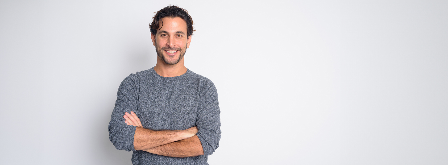 The image shows a man standing against a white background with his arms crossed, wearing a dark-colored top.