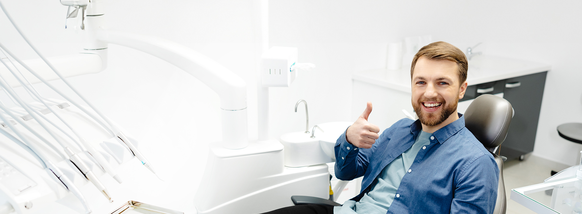 The image features a man sitting at a dental chair with his thumbs up, wearing a blue shirt, and smiling towards the camera.