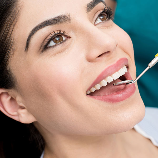 The image shows a woman with dark hair smiling broadly while holding a dental mirror up to her mouth, displaying a bright smile with a clean upper front tooth.